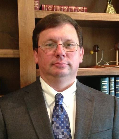 A man in a suit and tie stands in front of a bookshelf