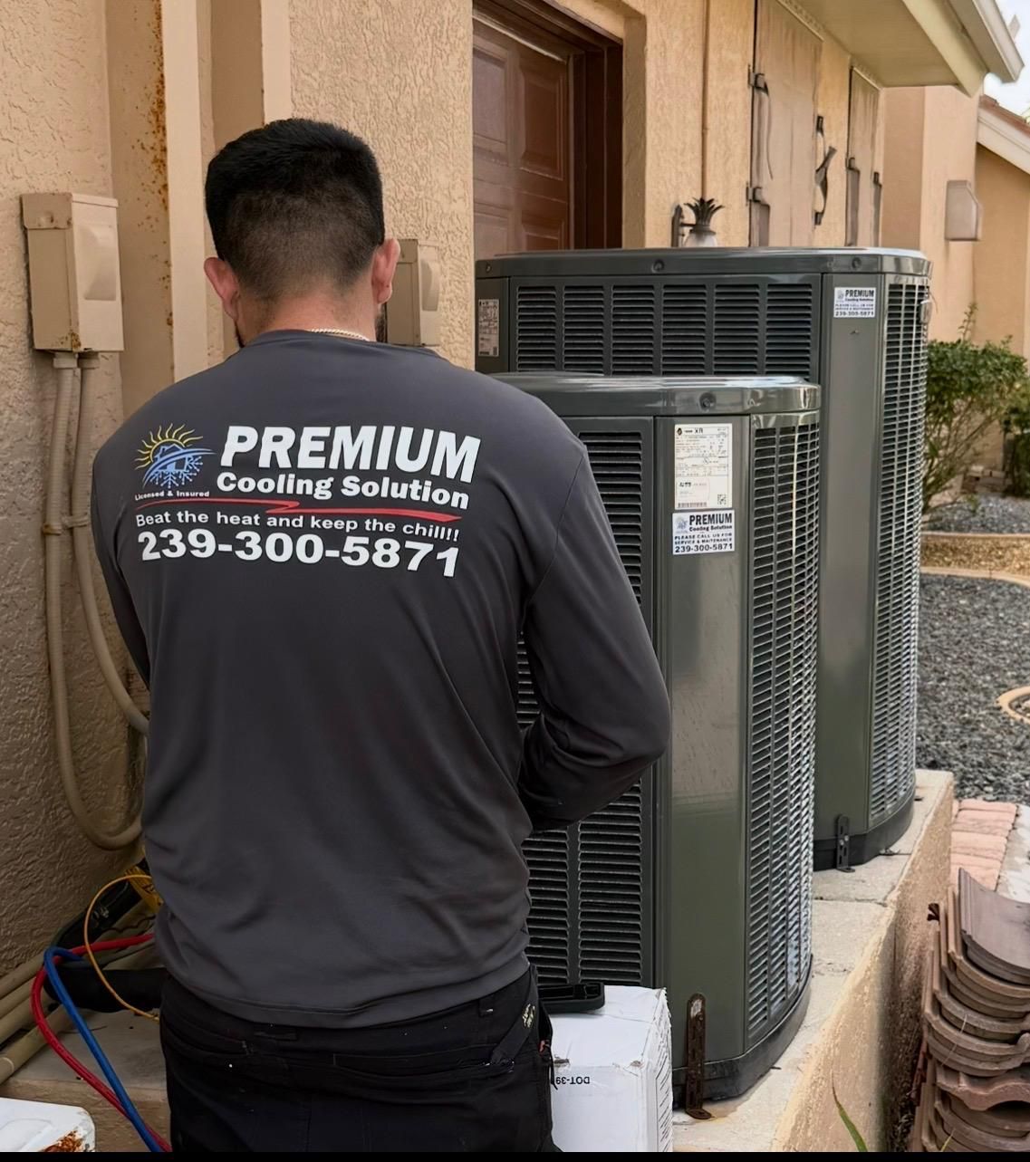 HVAC technician in gray shirt, working on air conditioning units outside a building. Contact info visible on shirt.