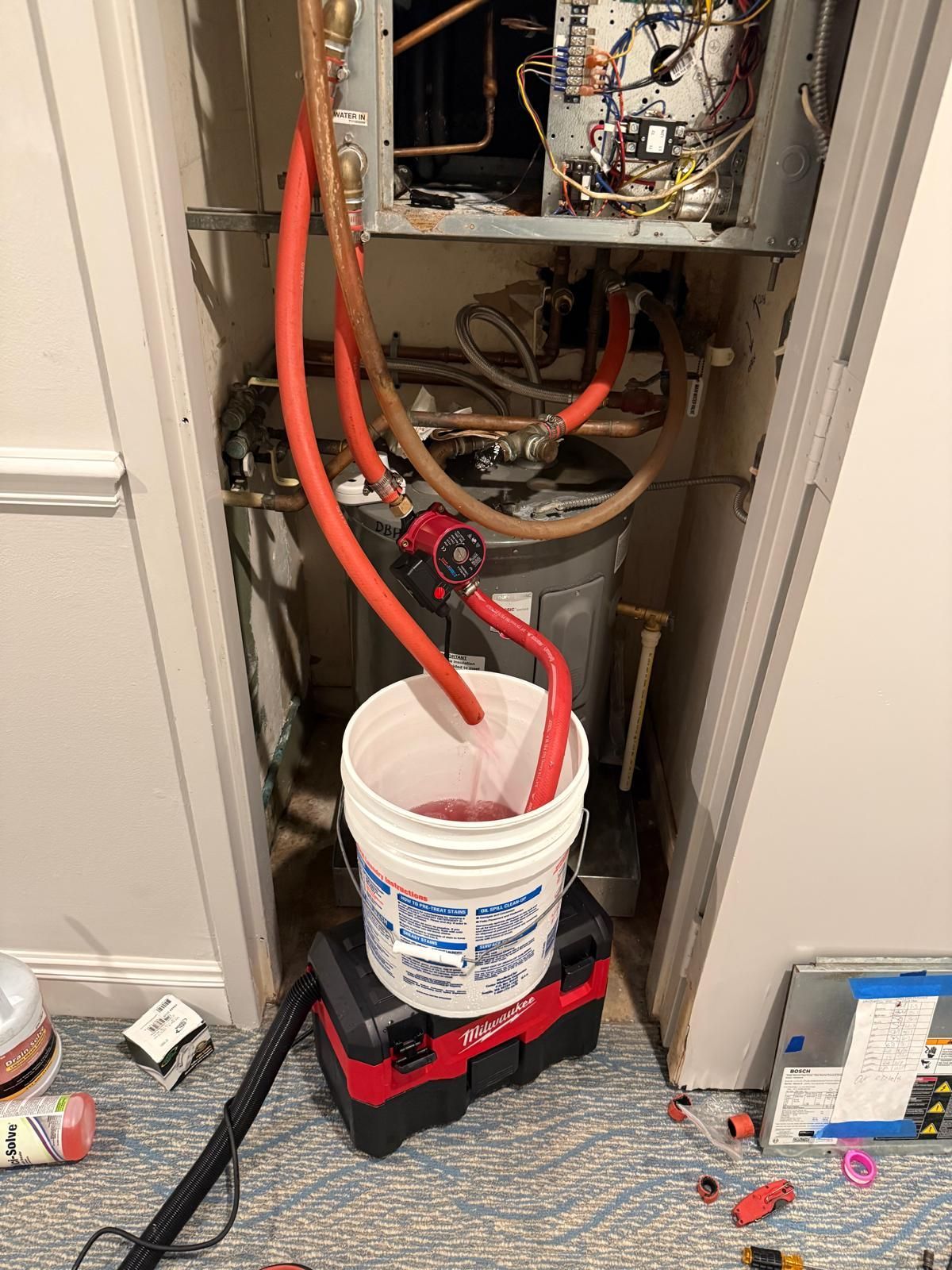 A water heater and components within a closet, with red hoses draining into a bucket atop a shop vacuum.