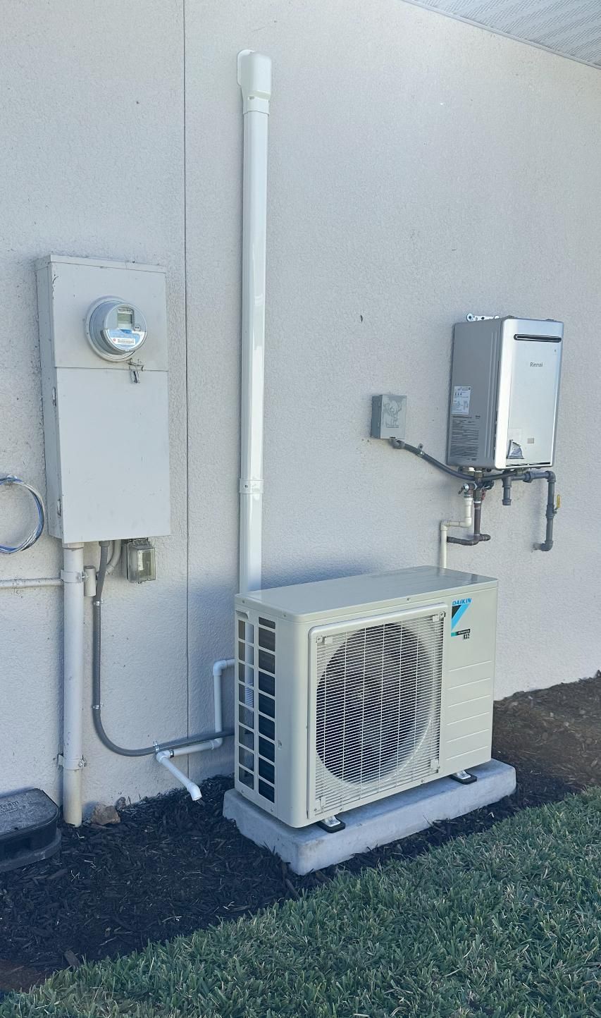 Outdoor utility equipment on a building wall: electrical box, air conditioner unit, water heater, and piping.