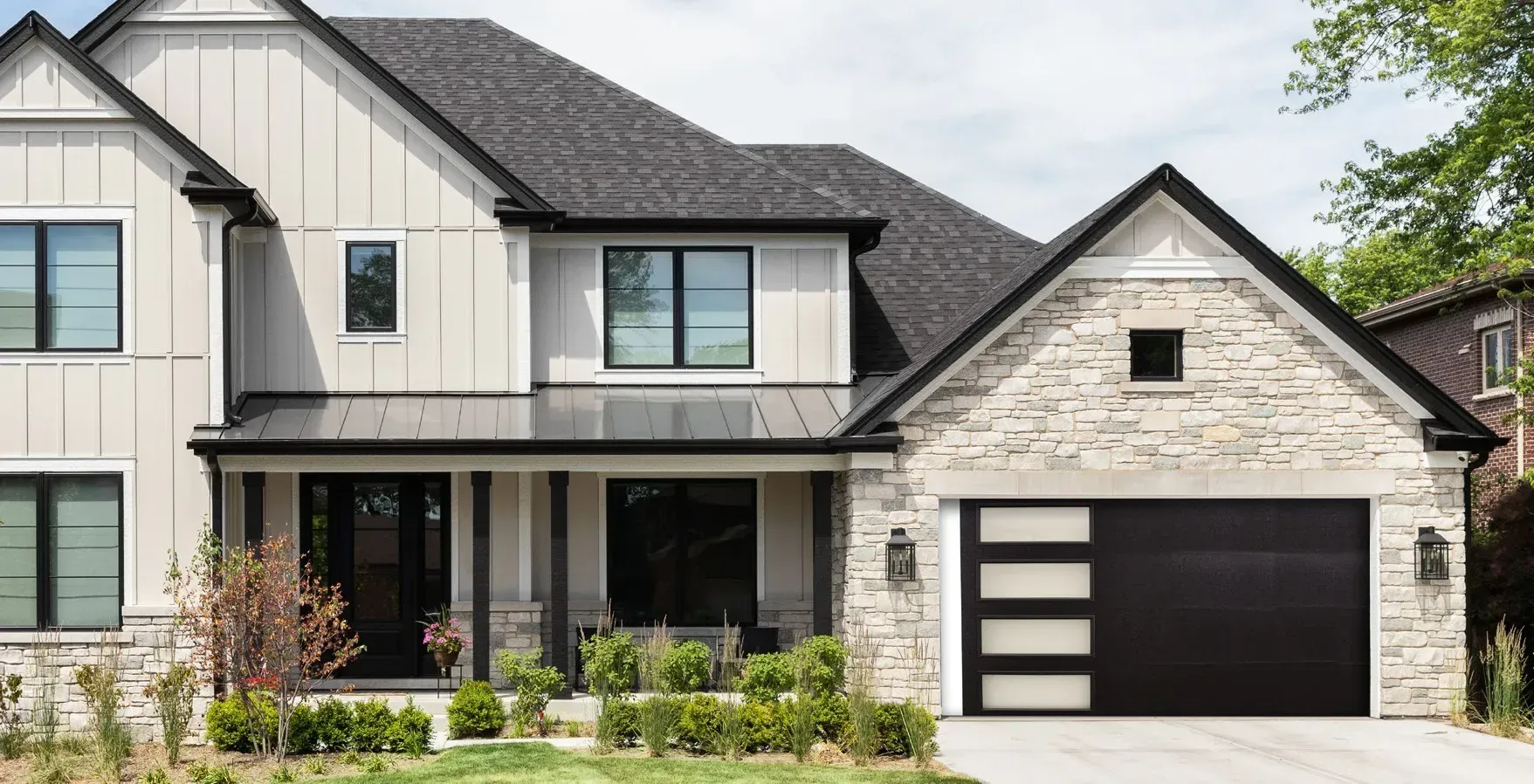 A modern home exterior with a light-colored garage door, a dark front door, stone walls, and a balcony with a black railing.