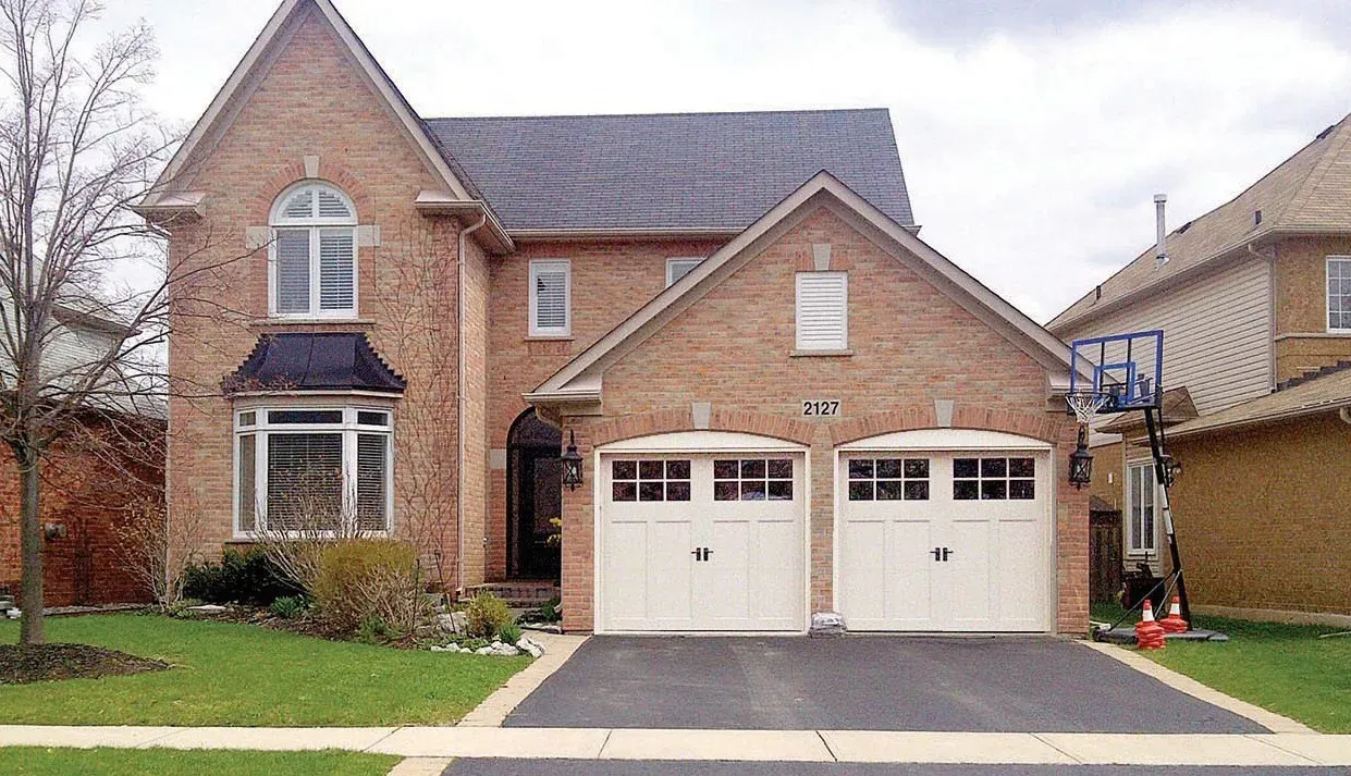 A white garage door with a pergola structure in front of a tan house with a tiled roof.