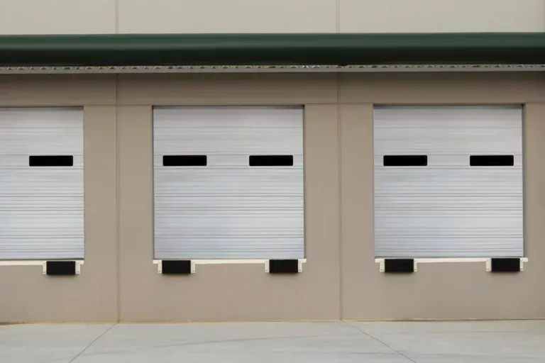 Three industrial roll-up warehouse loading dock doors with black window inserts and rubber bumpers on a beige exterior.
