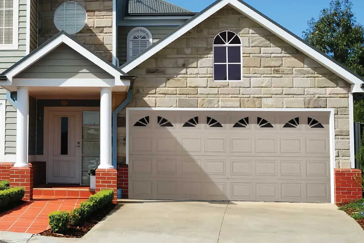 A beige, paneled garage door with decorative arched windows above, attached to a stone-faced house with a tiled entryway.
