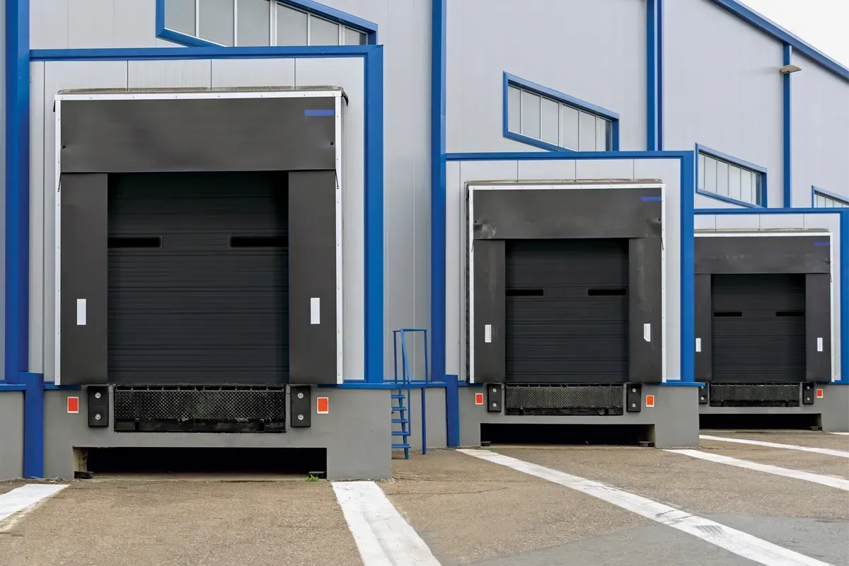 Three loading dock bays with closed black roll-up doors on a grey and blue warehouse building exterior.