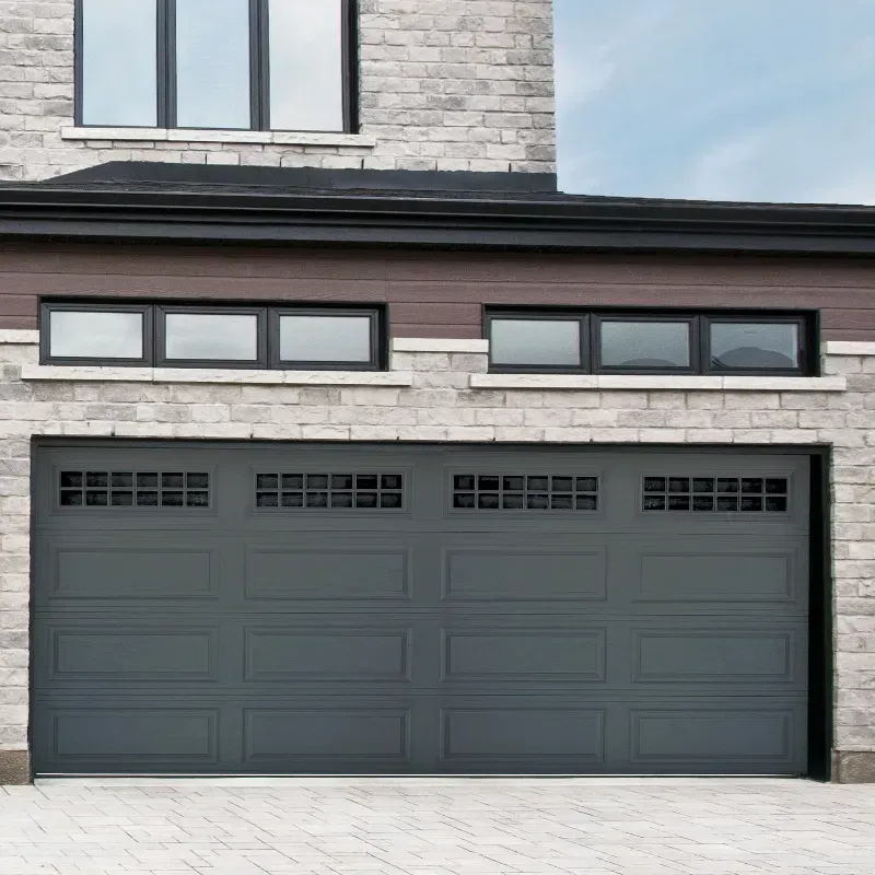A dark grey, four-panel residential garage door with rectangular window inserts, set against a light stone exterior wall.