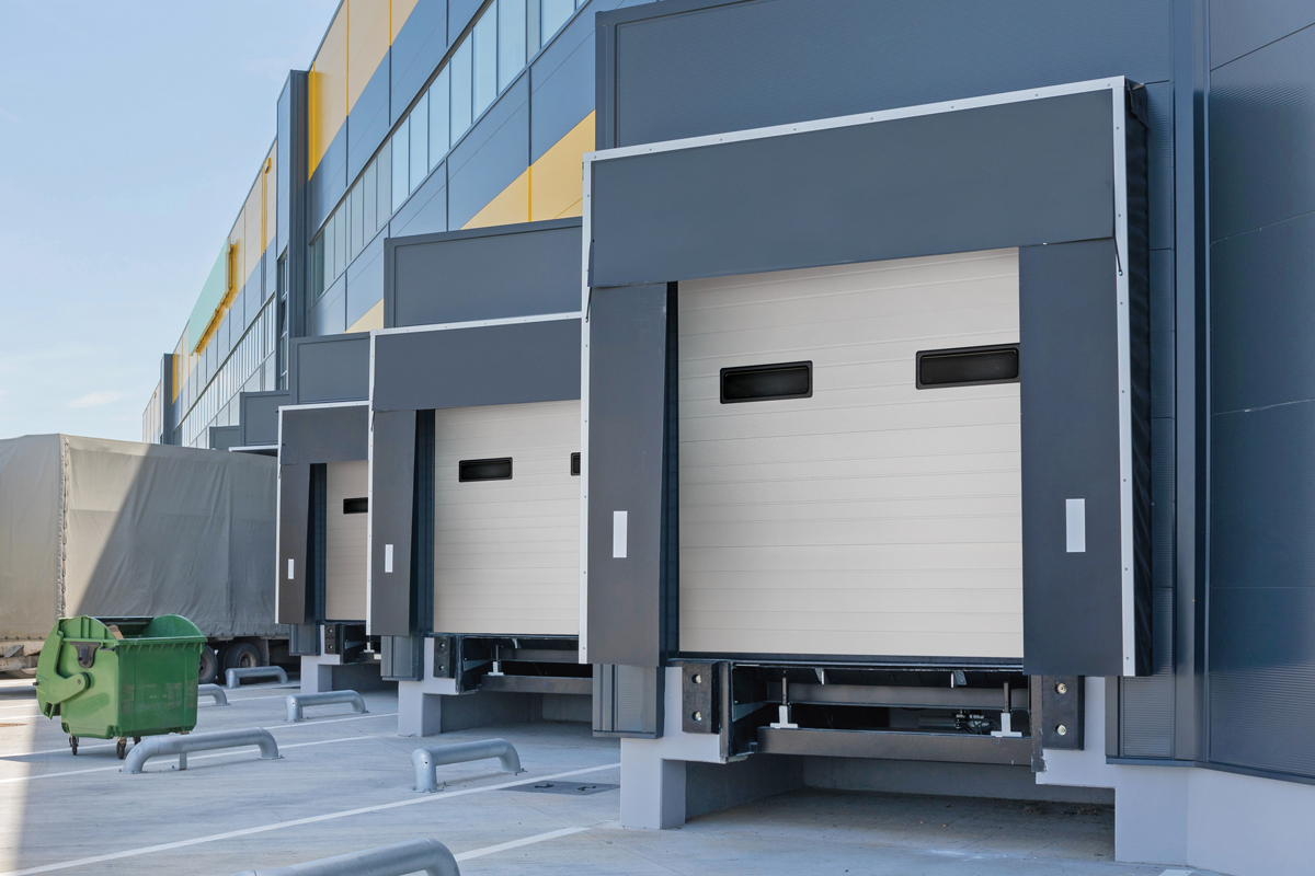 A row of industrial loading docks with roll-up beige doors and gray steel framing outside a modern warehouse facility.