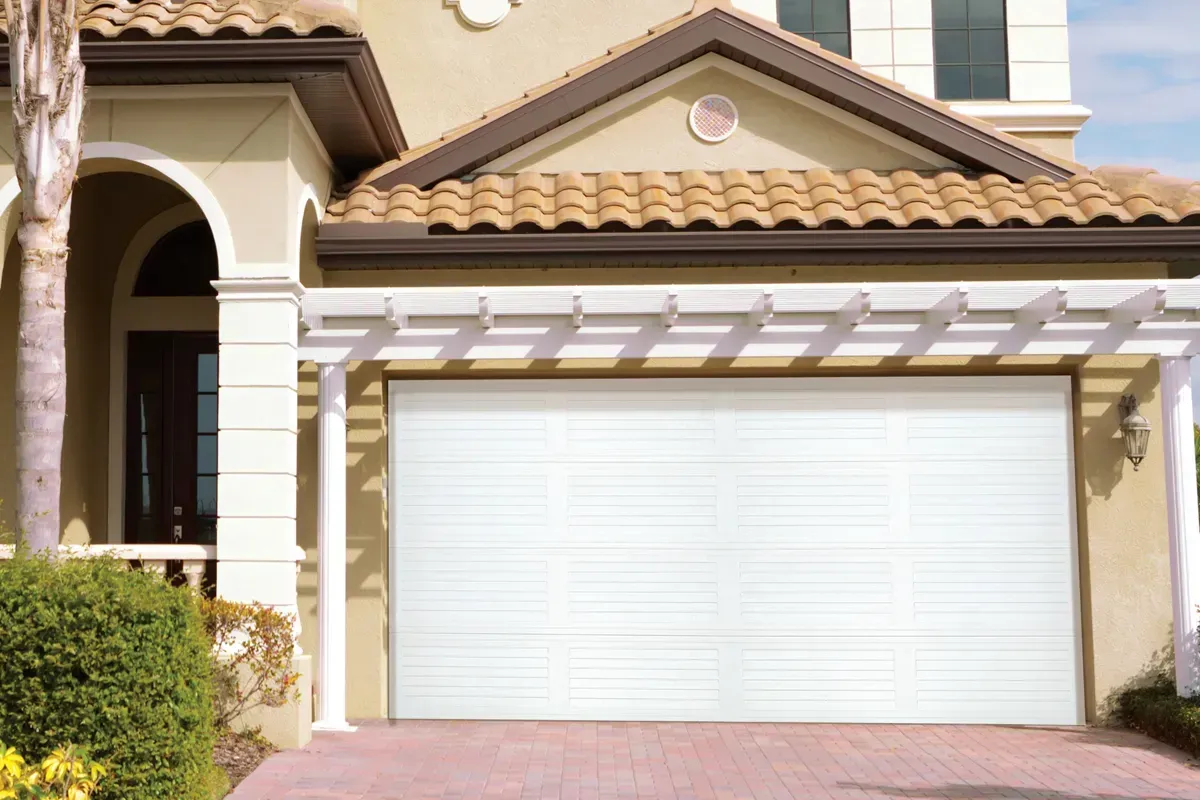 A white garage door with horizontal panels, beneath a wooden trellis, attached to a tan stucco house with a tiled roof.