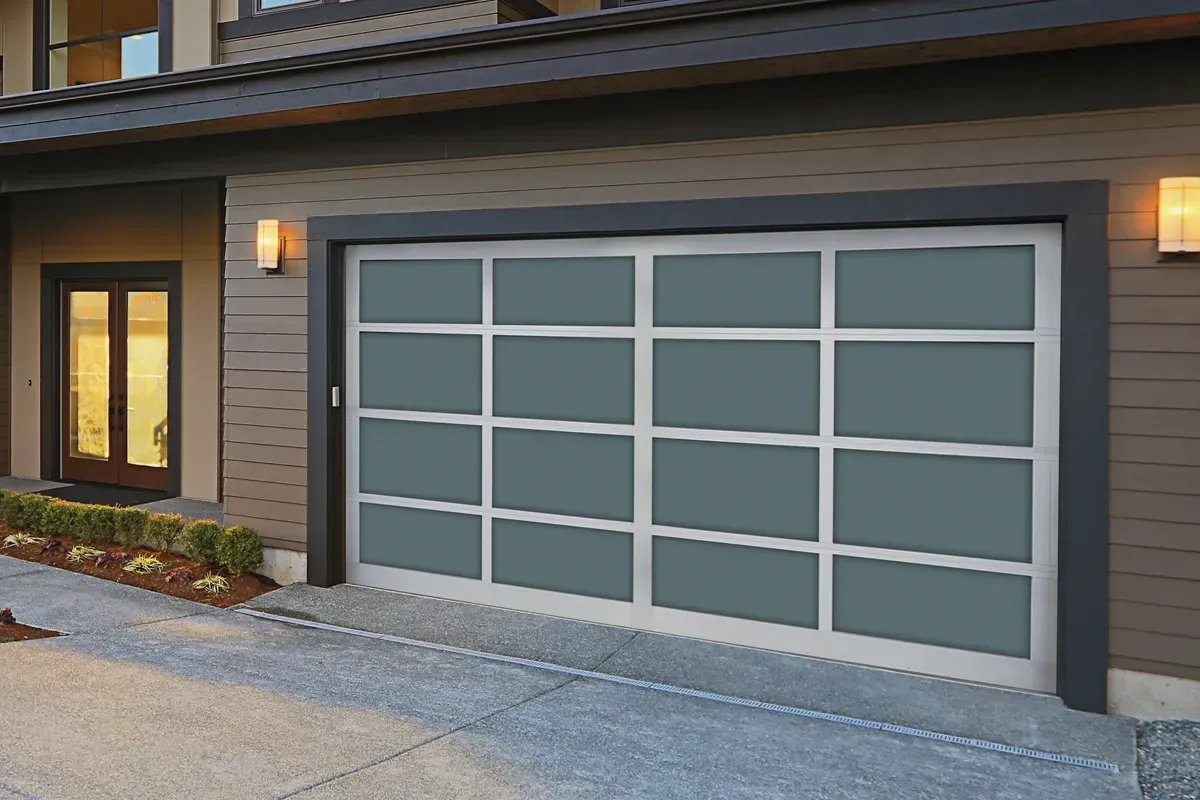 Modern residential garage door with silver frames and rectangular, opaque blue-grey glass panels on a dark-paneled house.