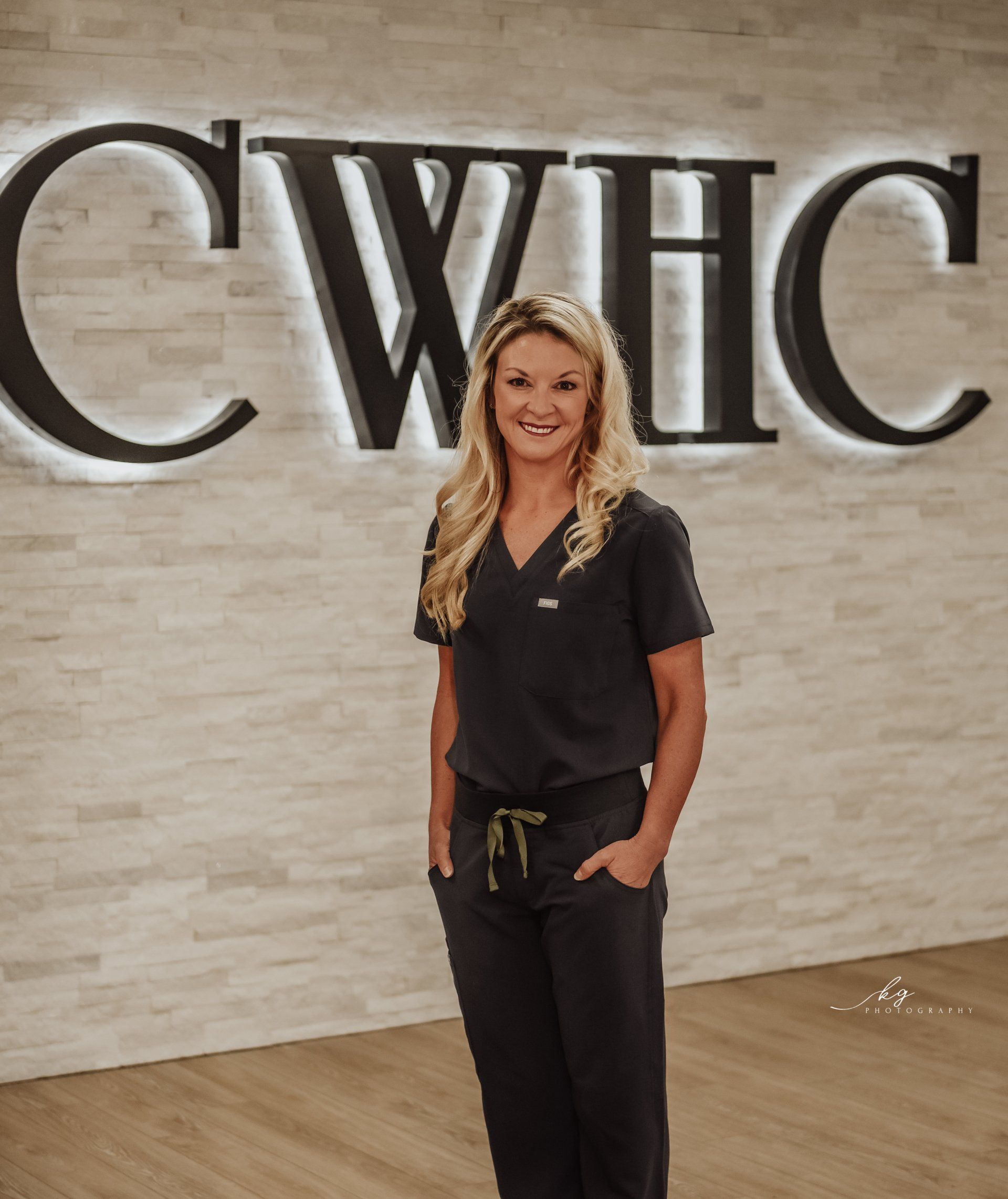 A smiling healthcare professional in dark charcoal scrubs stands in front of a white wall with backlit CWHC lettering.