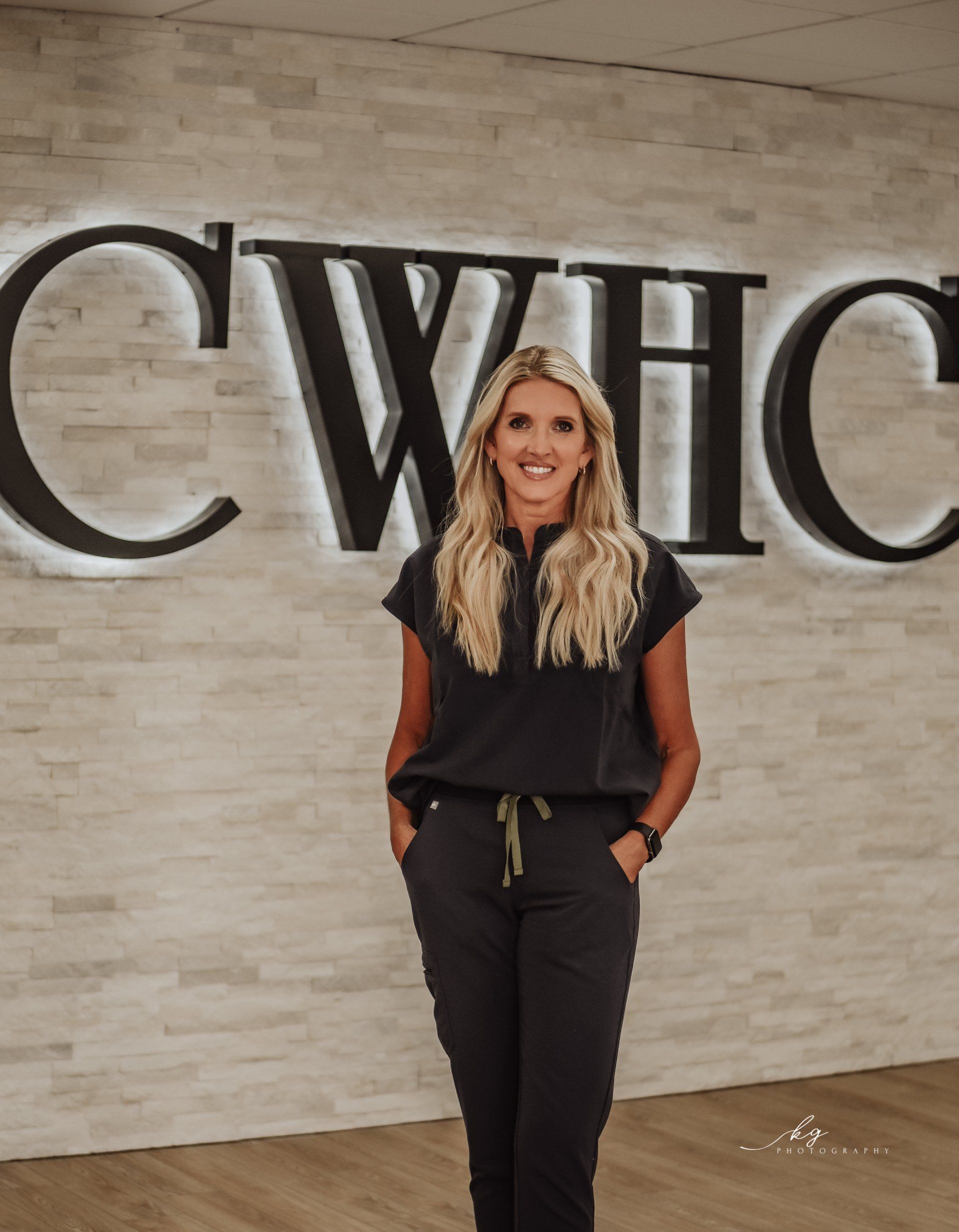 A smiling person with long blonde hair, wearing dark scrubs, stands in front of a stone wall featuring the letters CWHC.