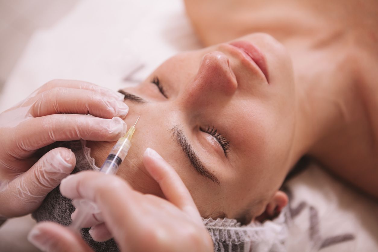A practitioner in medical gloves administers a facial injection to a person resting on a treatment table.