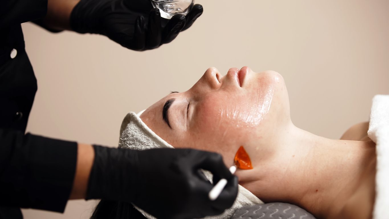 A person in black gloves uses an orange spatula to apply a skincare treatment to a client's face.