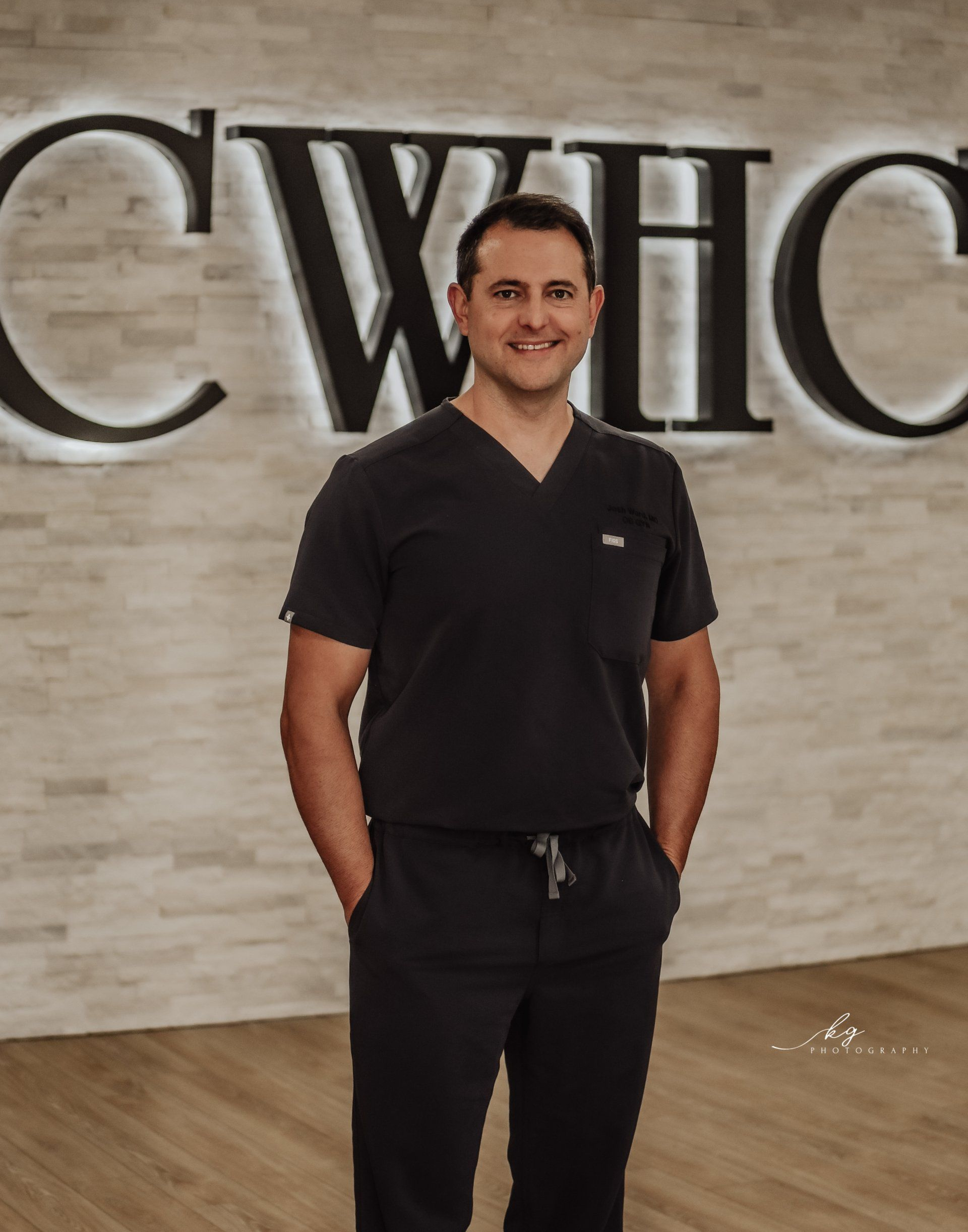 A healthcare professional in dark scrubs smiling in front of a wall with a backlit 
