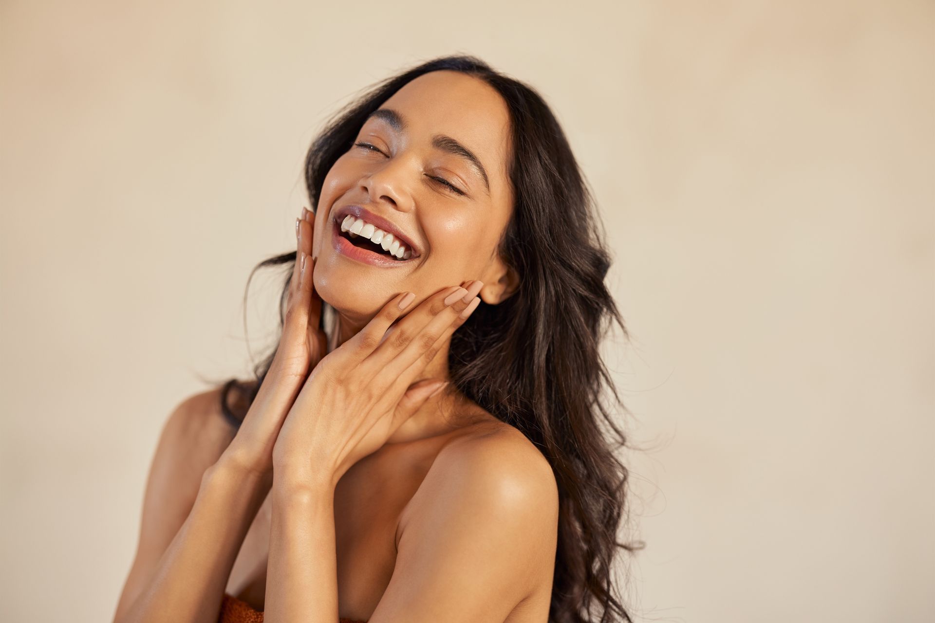 A person with long, dark hair touching their glowing, radiant face while laughing.