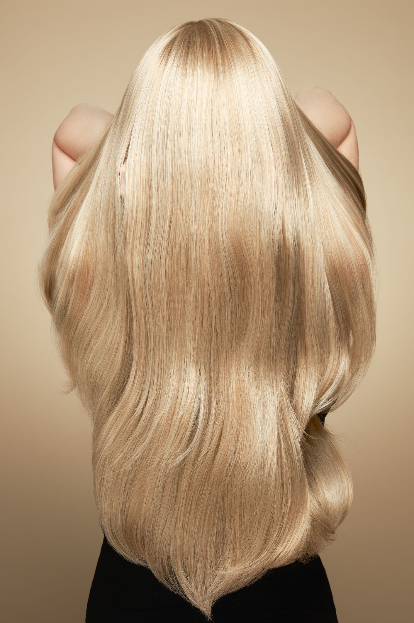 A person seen from behind, holding long, thick, blonde wavy hair that fans out over their upper back against a tan background.