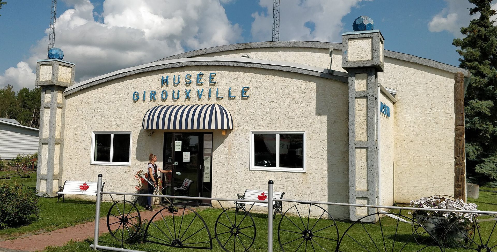 The Girouxville Museum, a light-colored building with a blue-striped awning, stands under a partly cloudy sky.