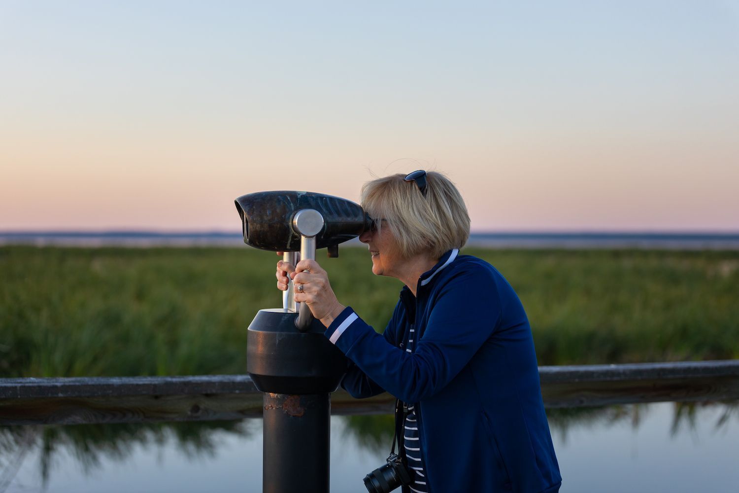 A person peers through a stationary viewing scope at a marshy landscape during sunset.