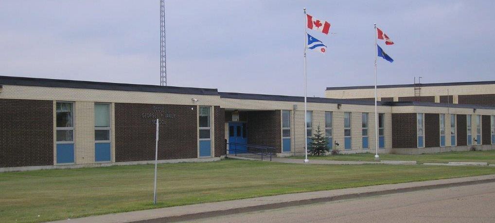A single-story school building with brown and beige panels, blue window frames, and two flagpoles flying Canadian flags.