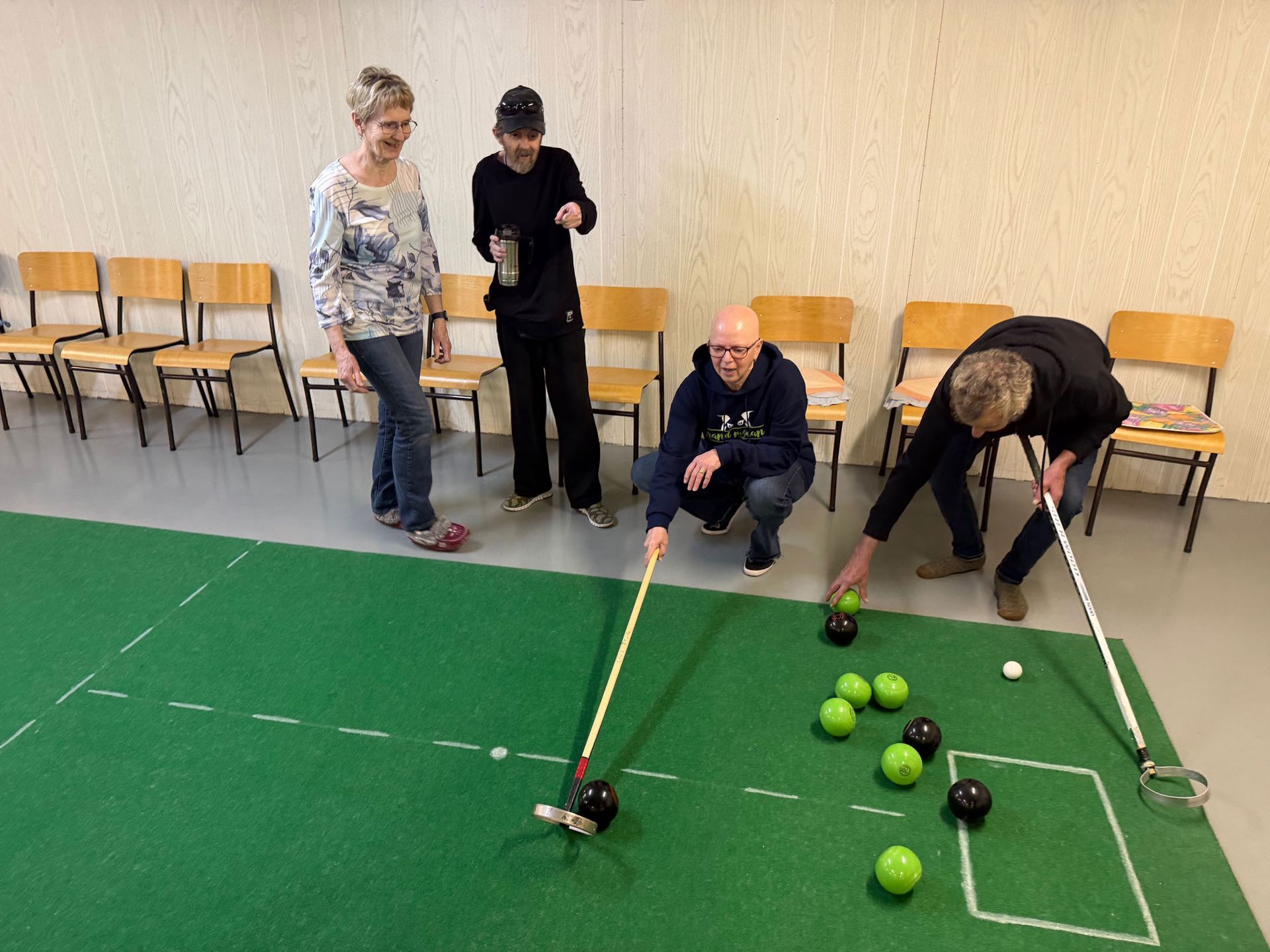 Four people playing indoor carpet bowls on a green mat in a room with a row of wooden chairs.