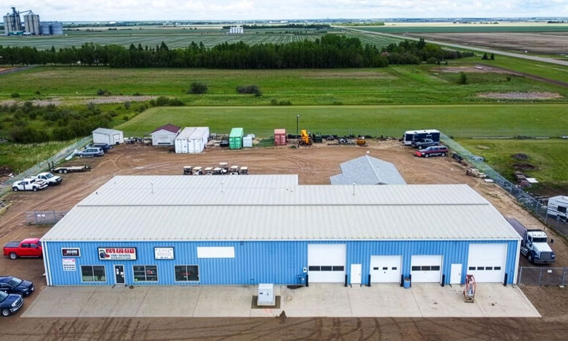 An aerial view of a blue, single-story commercial metal building with four white garage doors, set in a rural landscape.