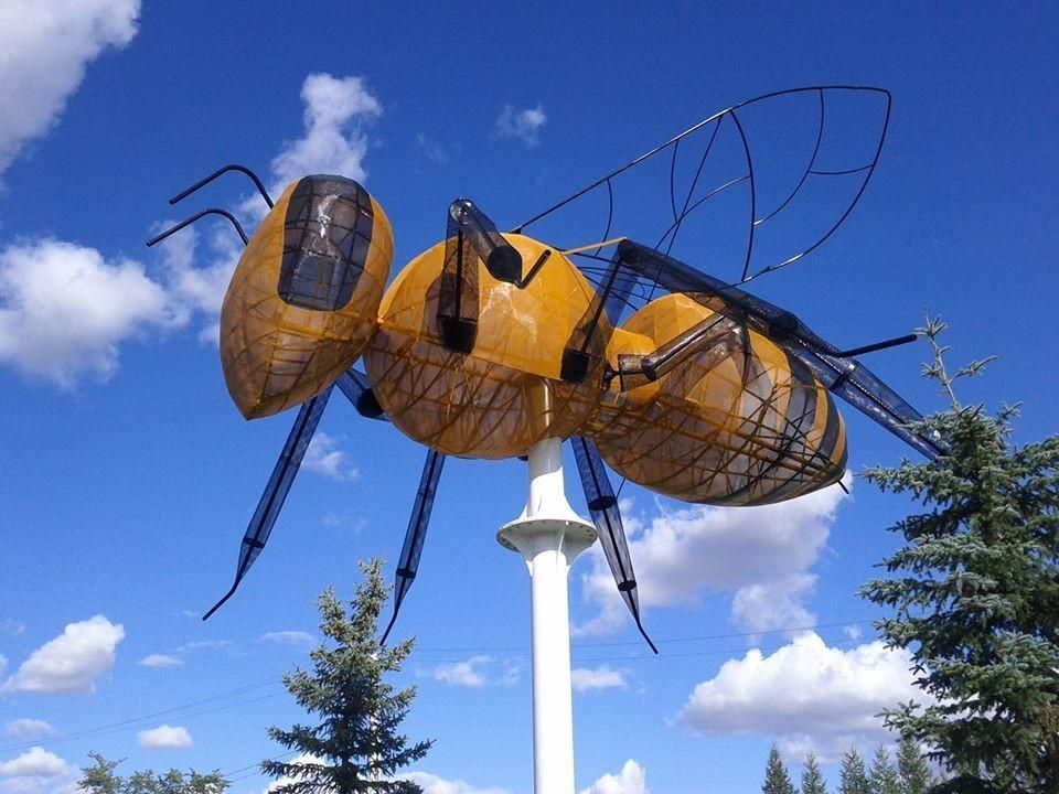 A large, yellow, mesh-constructed sculpture of a bee mounted on a white pole against a blue, cloudy sky.