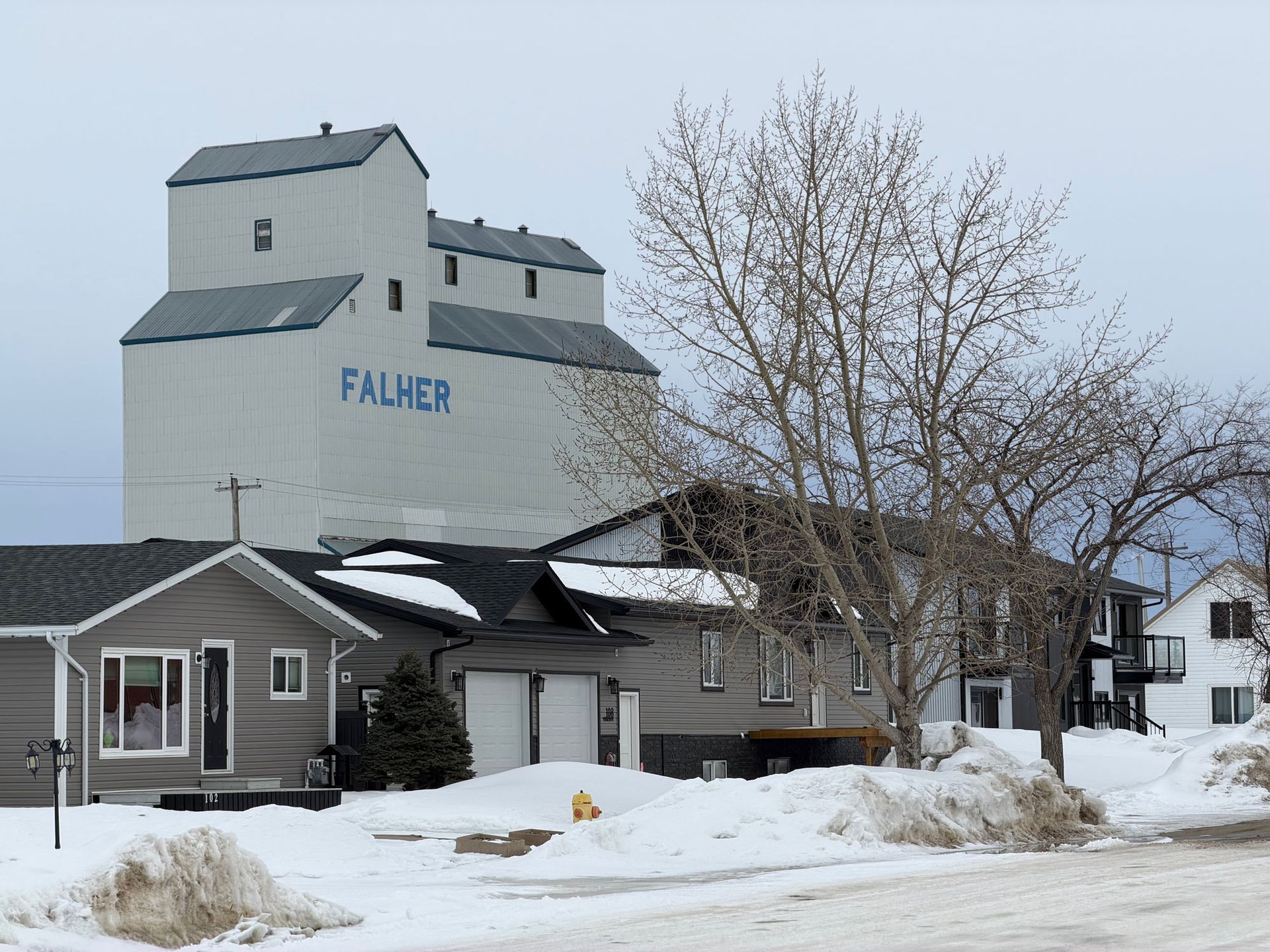 A large gray Falher grain elevator towers over residential houses and snow-covered ground on a cloudy day.