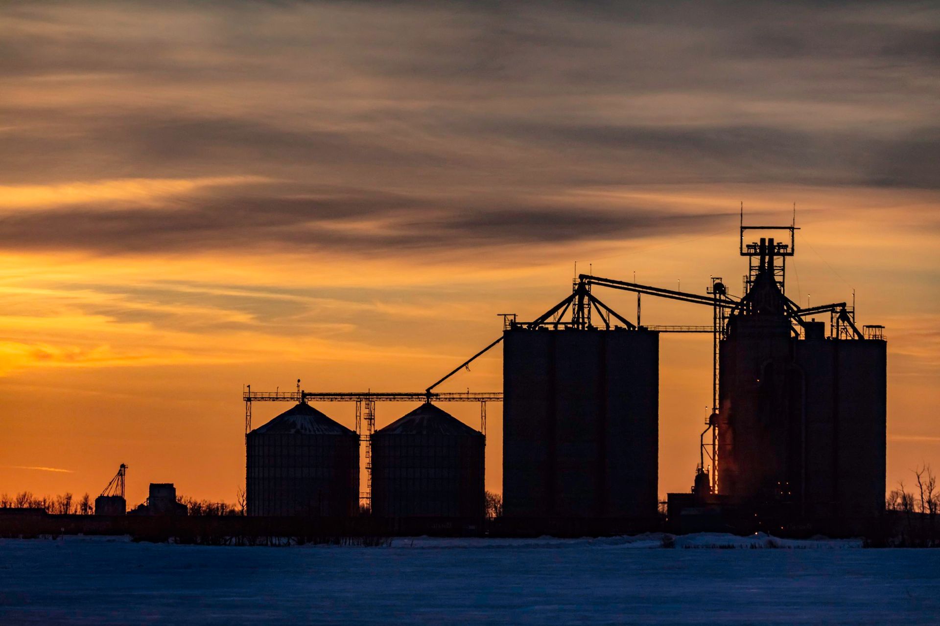 A silhouette of grain silos against a vibrant orange and purple sunset over a snow-covered field.