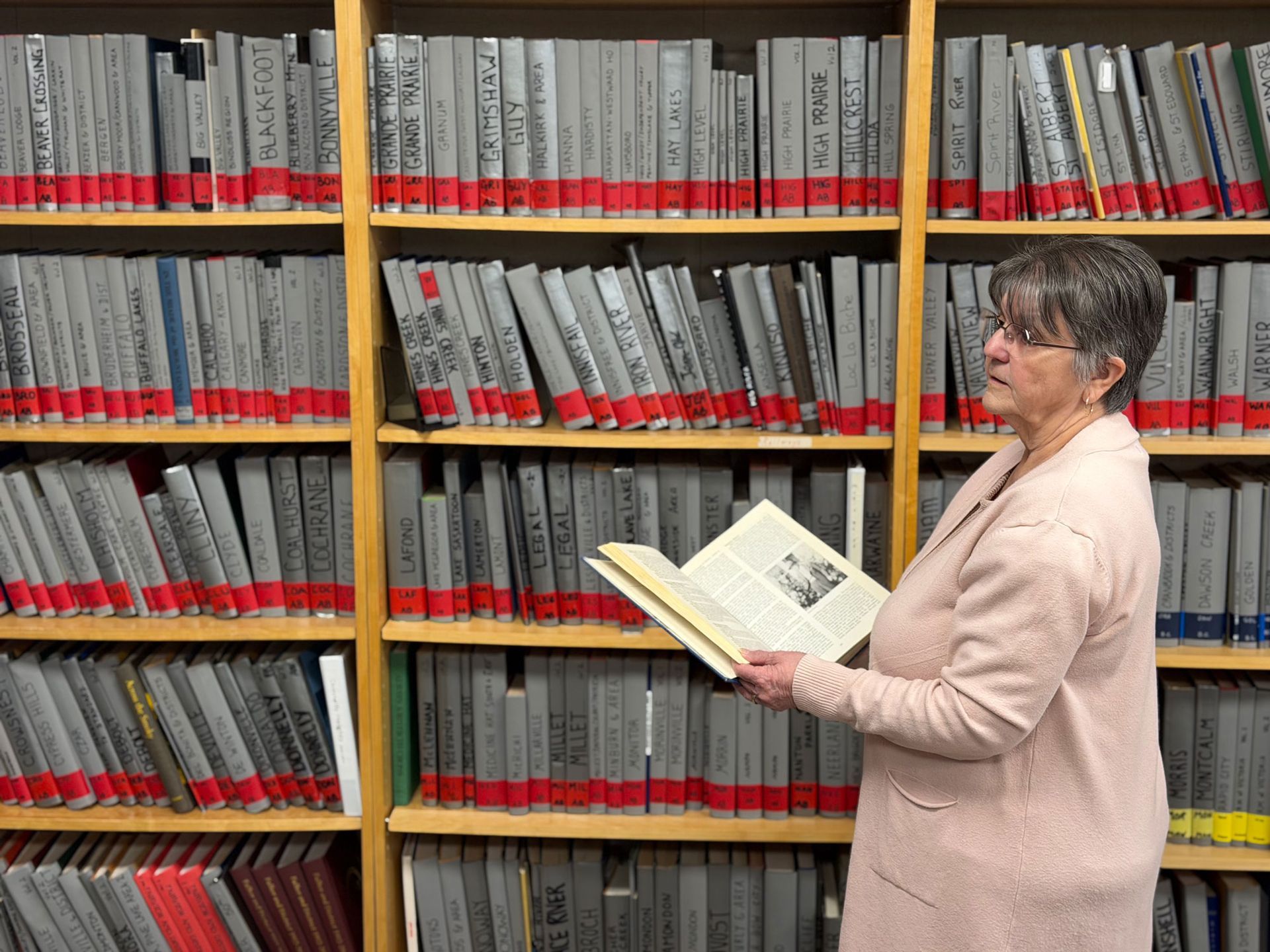 A person standing in front of library shelves filled with grey-spined books, holding and reading an open book.