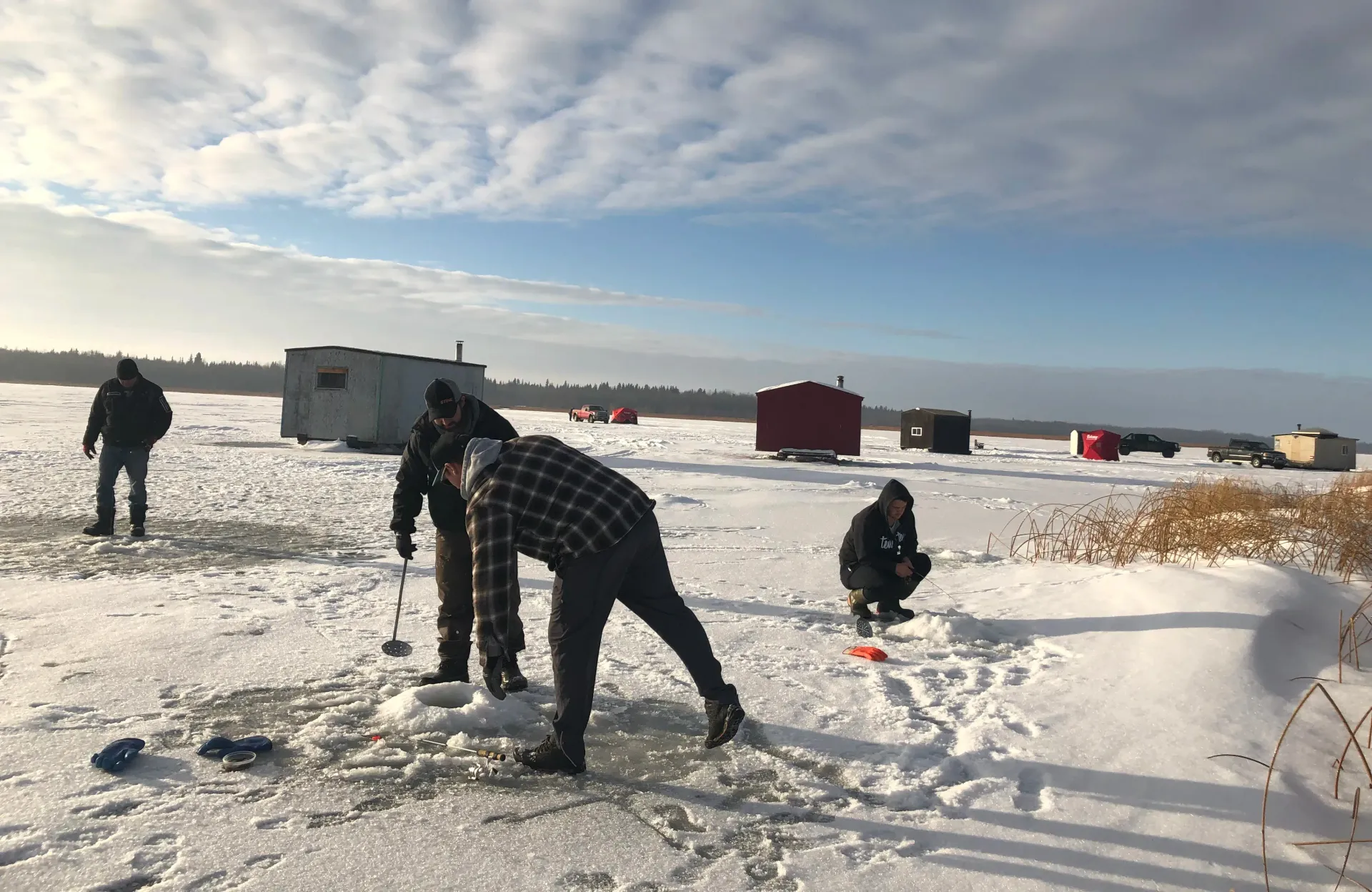People ice fishing on a snowy, frozen lake near several small fishing shelters under a partly cloudy blue sky.