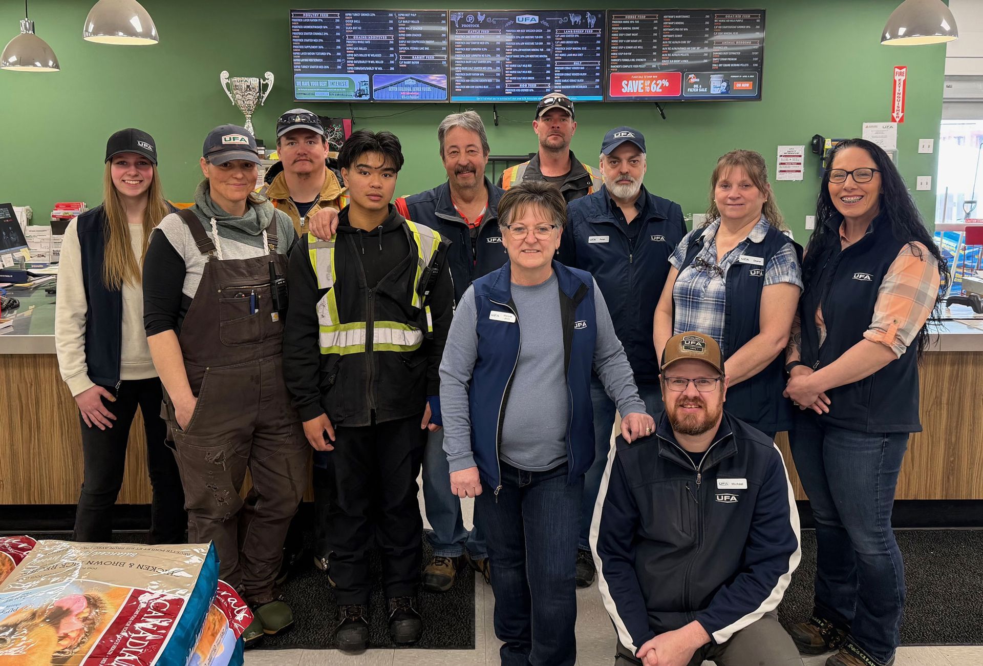 A group of eleven staff members in uniform stand smiling inside a workplace with a menu board on the green wall behind.