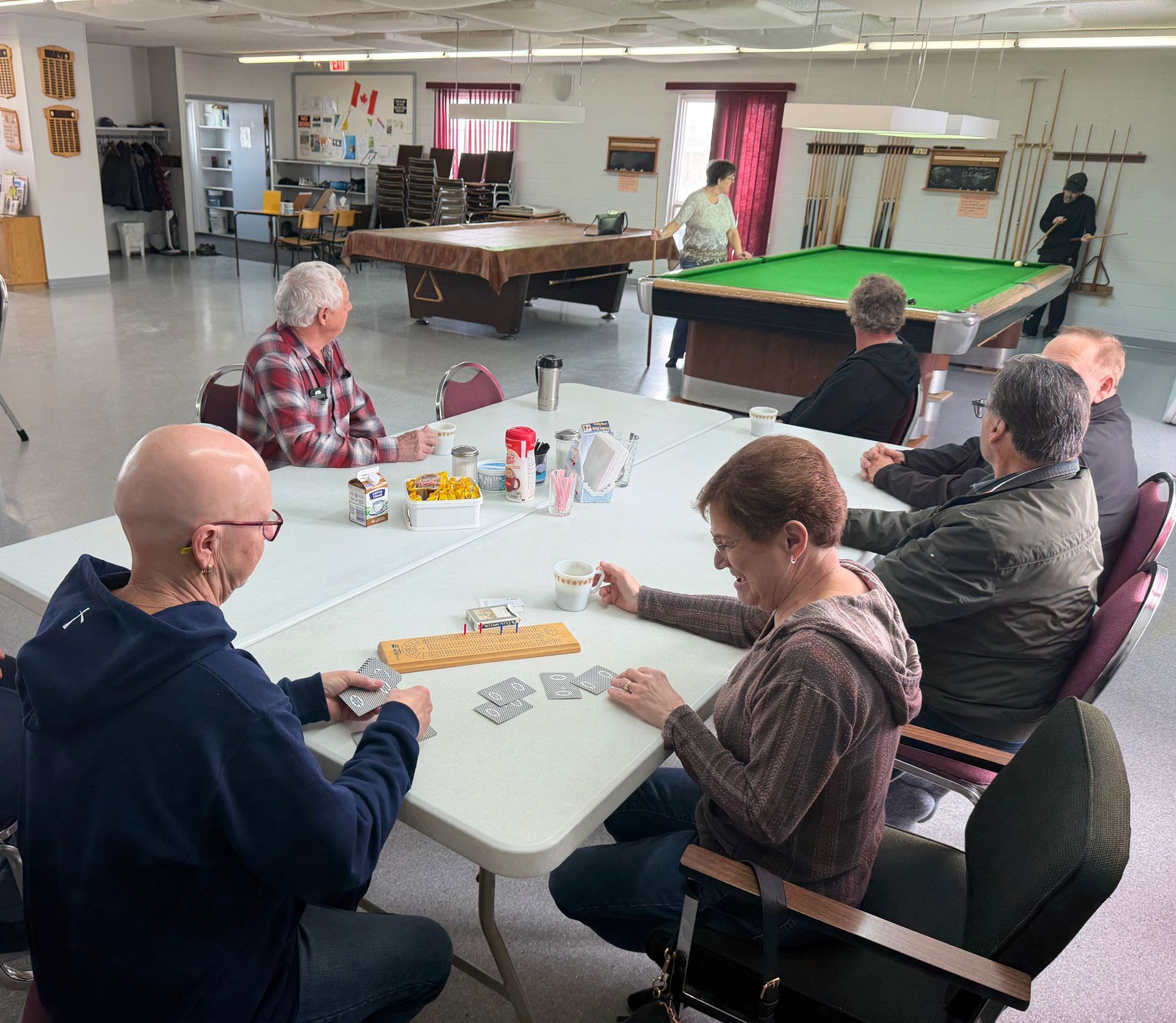 People play cards around a table in a community room with billiard tables in the background.