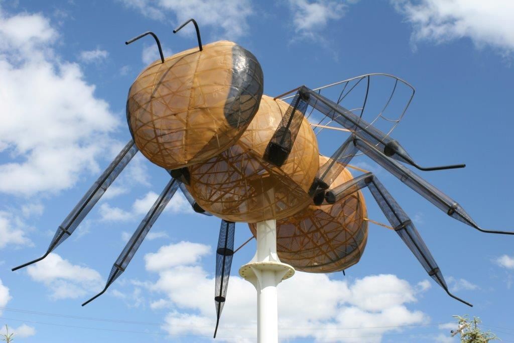 A large, tan-colored bee sculpture mounted on a white pole against a blue, cloudy sky.