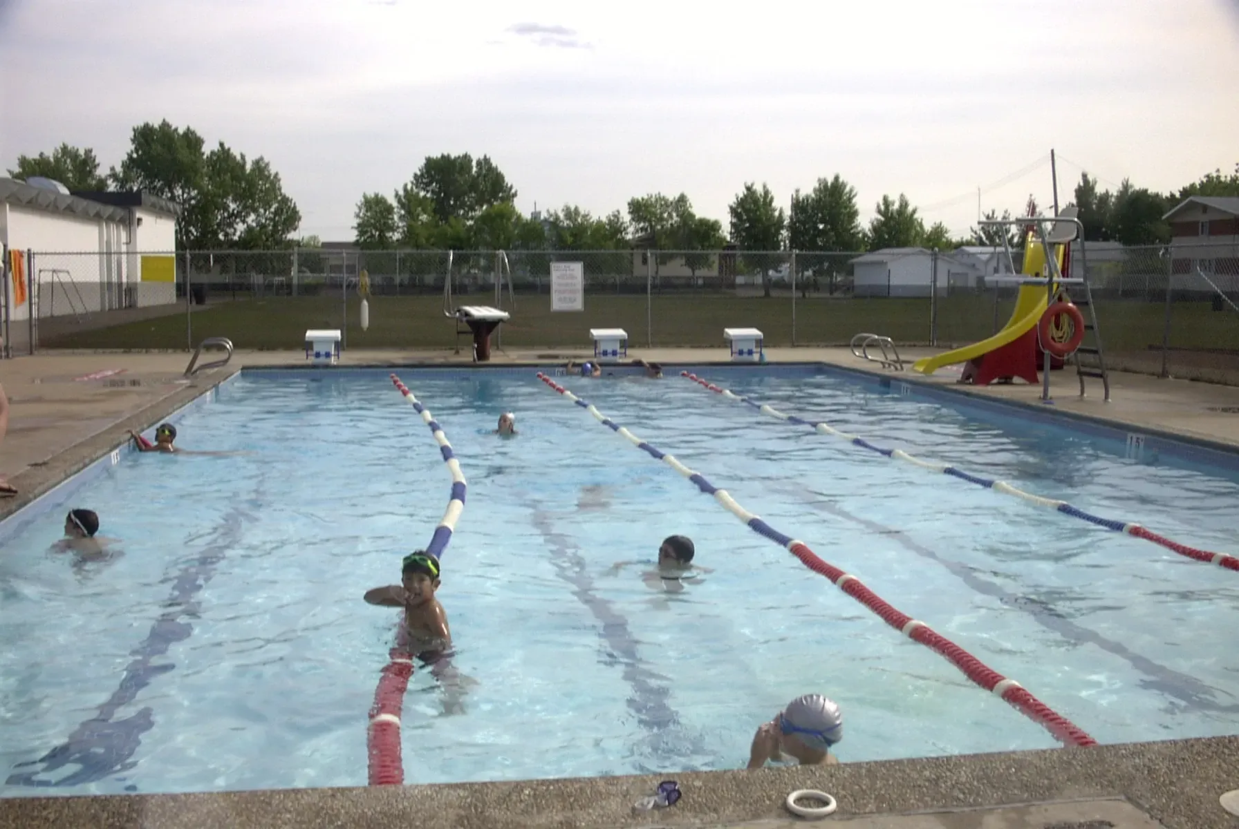 Children swim in an outdoor lap pool with lane dividers, a slide, and starting blocks under a bright, cloudy sky.