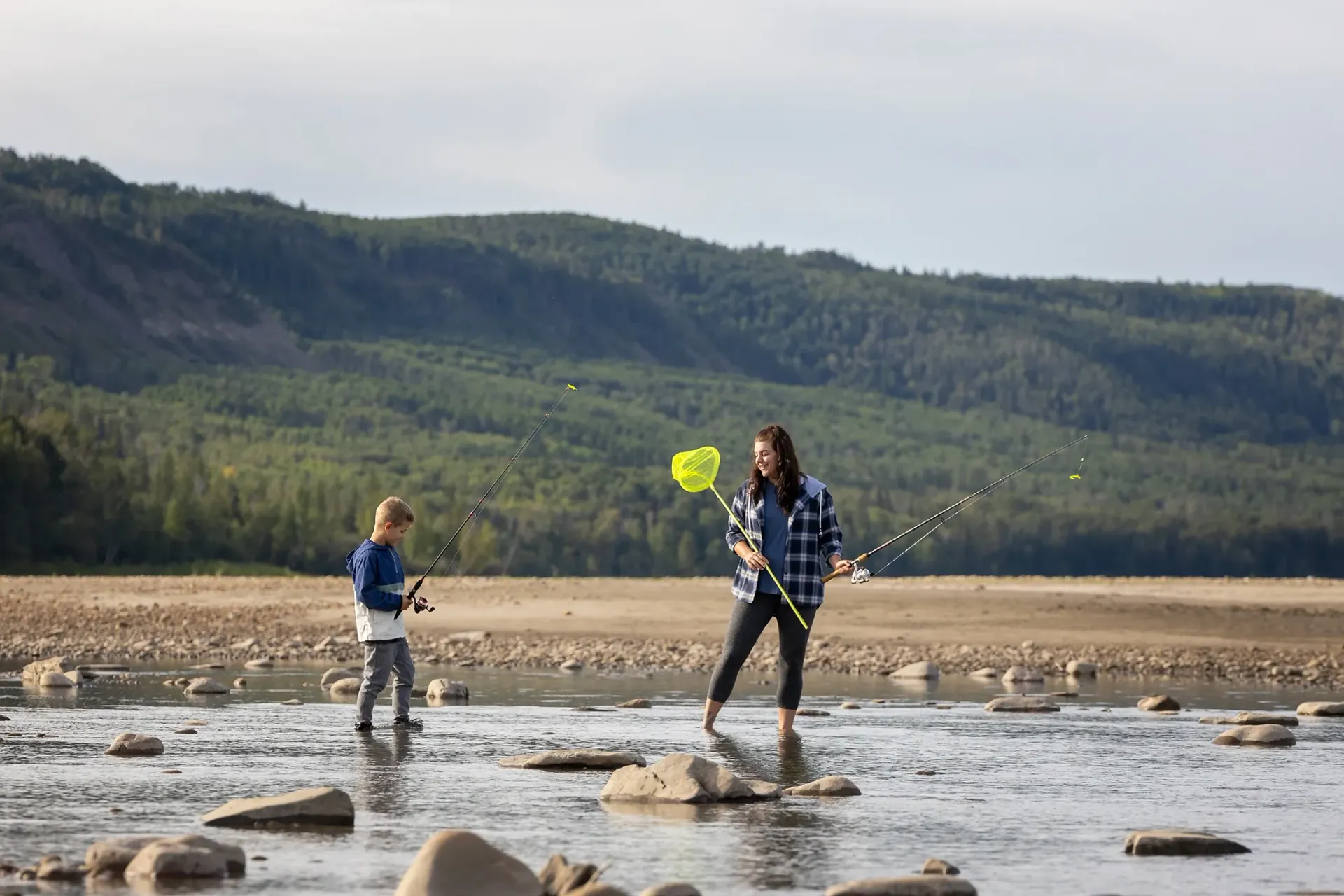 An adult and child wade through a shallow, rocky river while fishing with a rod and a neon yellow net.