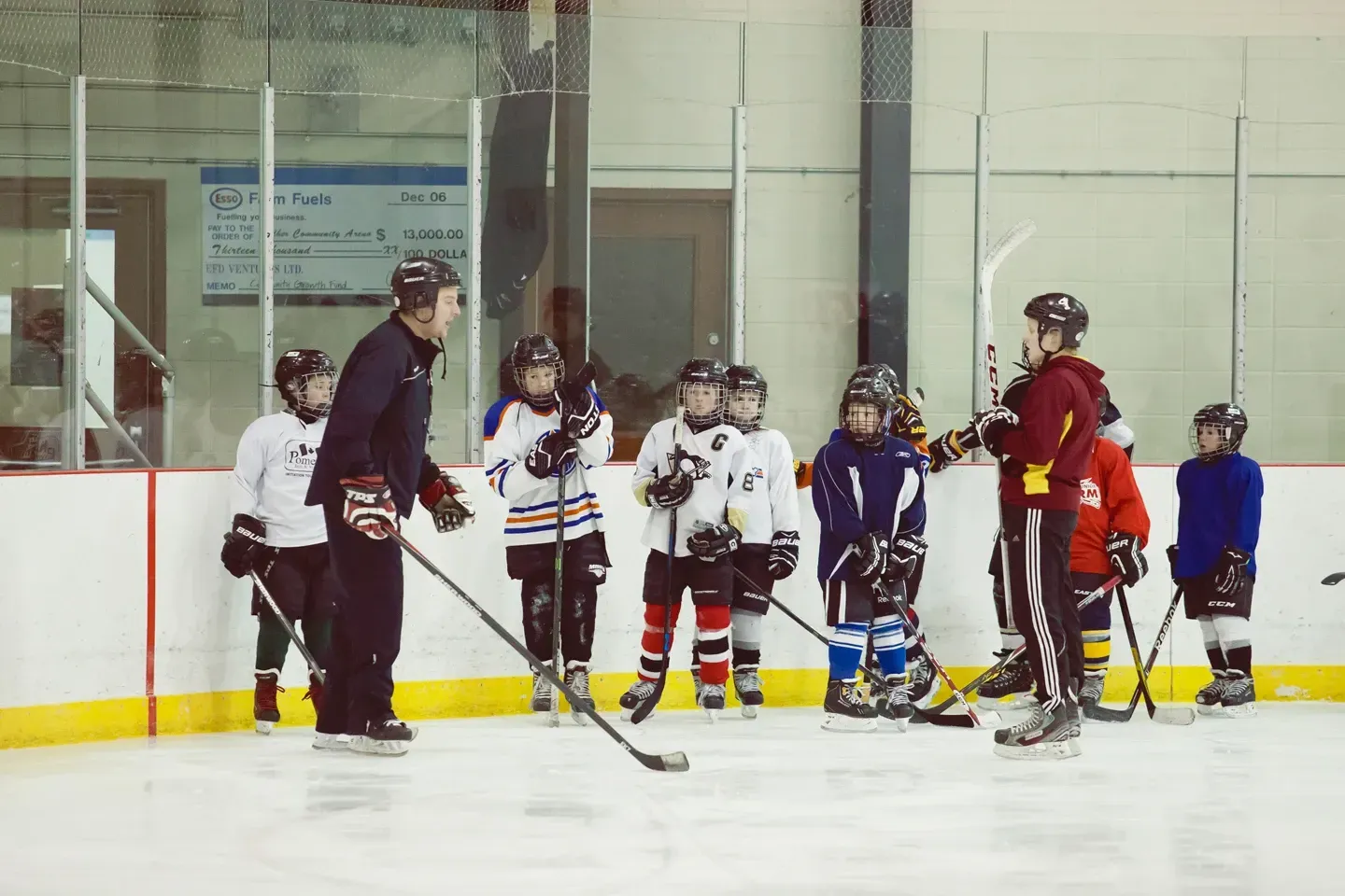A coach instructs a group of young hockey players wearing gear and skates on an indoor ice rink.