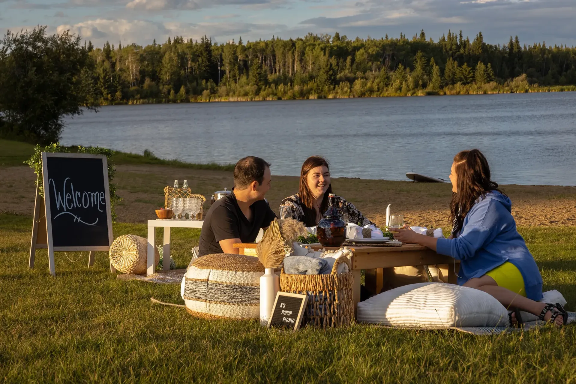 Three people enjoy a picnic on a grassy lakeshore next to a welcome sign and a small table at sunset.