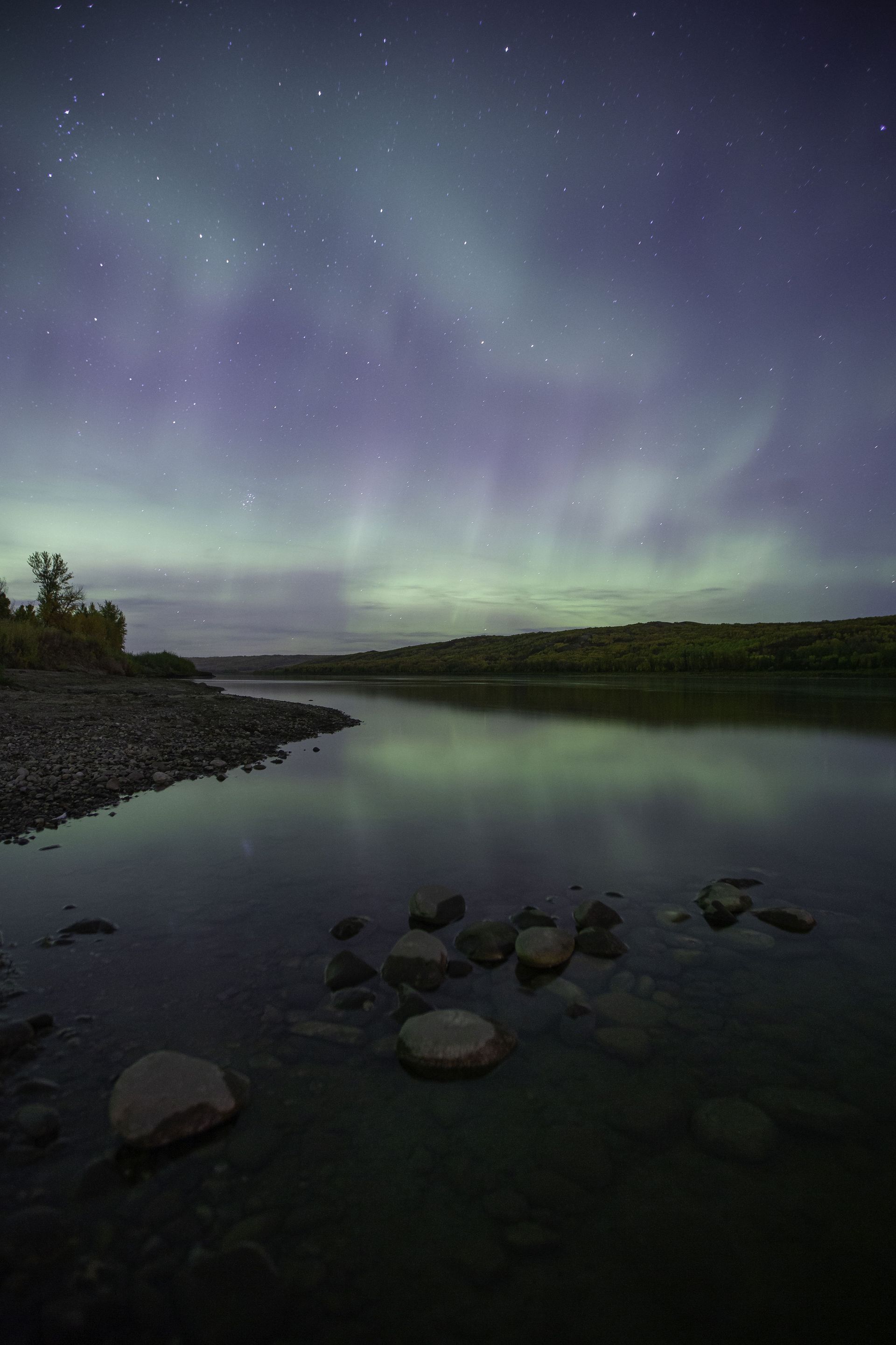 Aurora borealis illuminates a night sky over a calm river with rocky shores.
