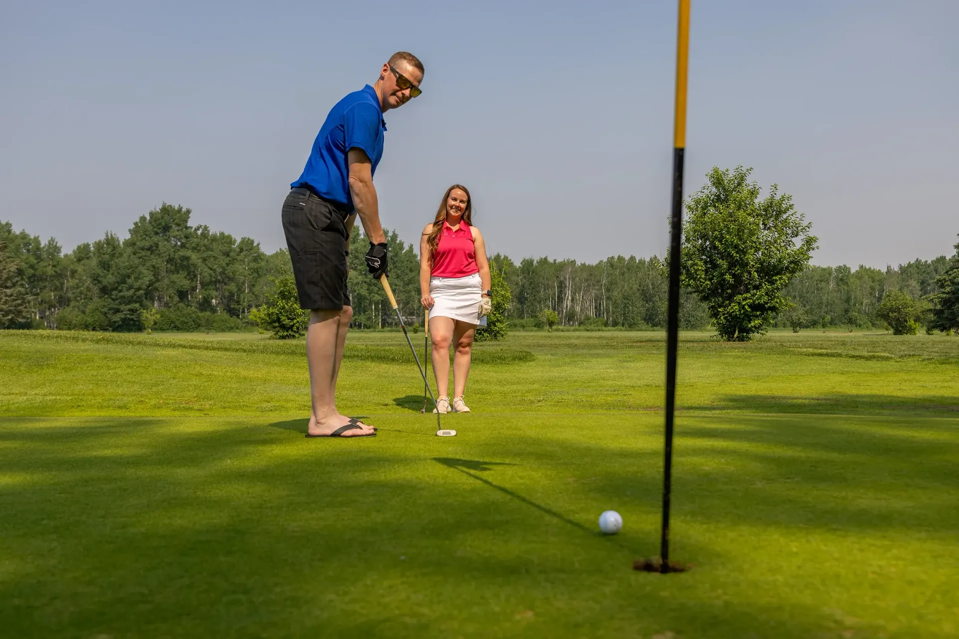 A golfer in a blue shirt prepares to putt on a green while a companion stands nearby in a sunny, wooded golf course setting.