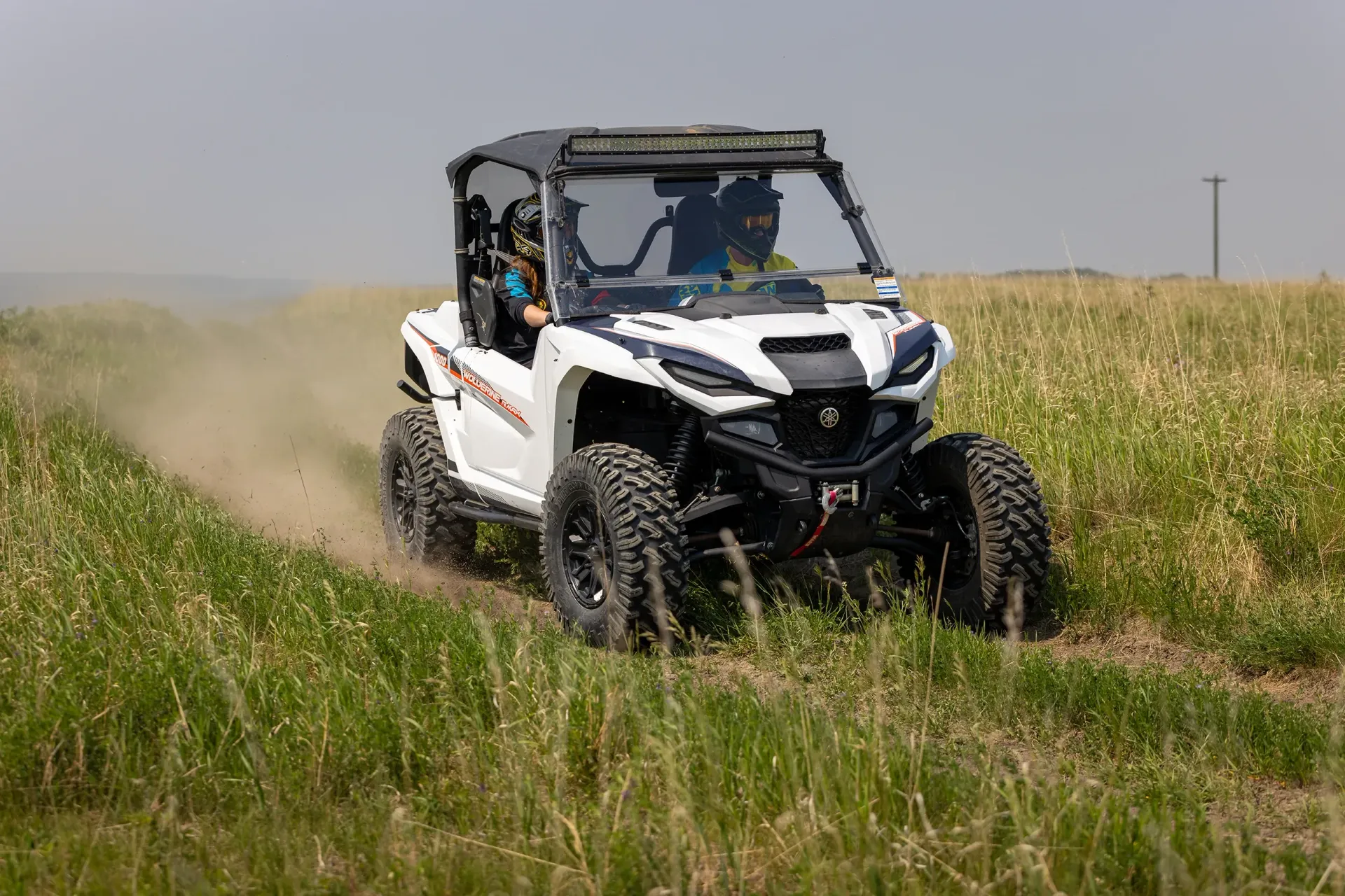 A white and black off-road utility vehicle driving through a tall, grassy field, kicking up dust.