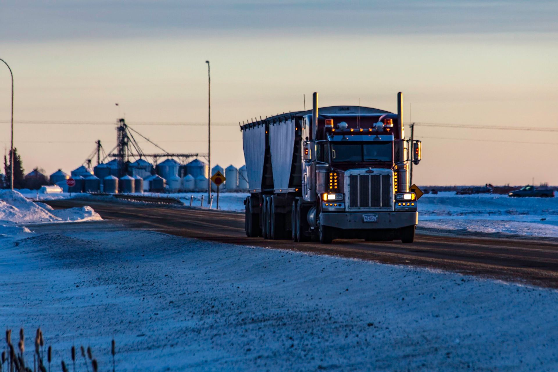 A semi-truck driving on a snow-covered road past a grain elevator during sunset.