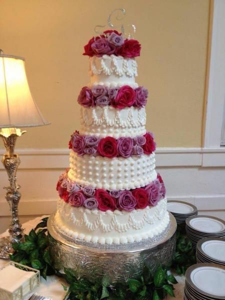 Tiered white wedding cake with pink and purple roses on a silver stand, surrounded by greenery.