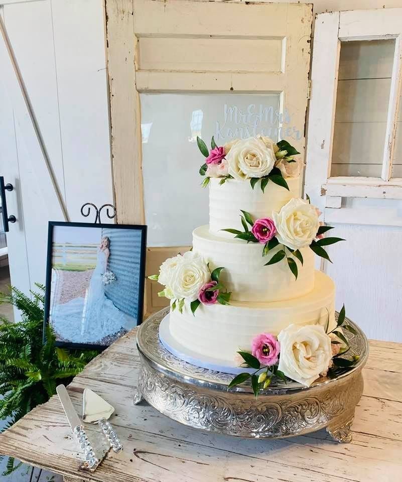 Four-tier white wedding cake with cream roses and pink accents on a rustic table indoors