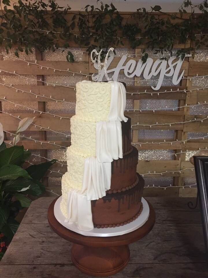 Tiered white-and-chocolate drip cake on a wooden stand, with a “Menggi” sign in the background.