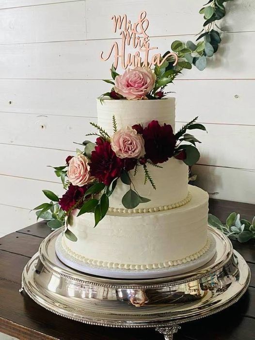 Three-tier white wedding cake with pink roses, greenery, and a gold topper on a silver tray