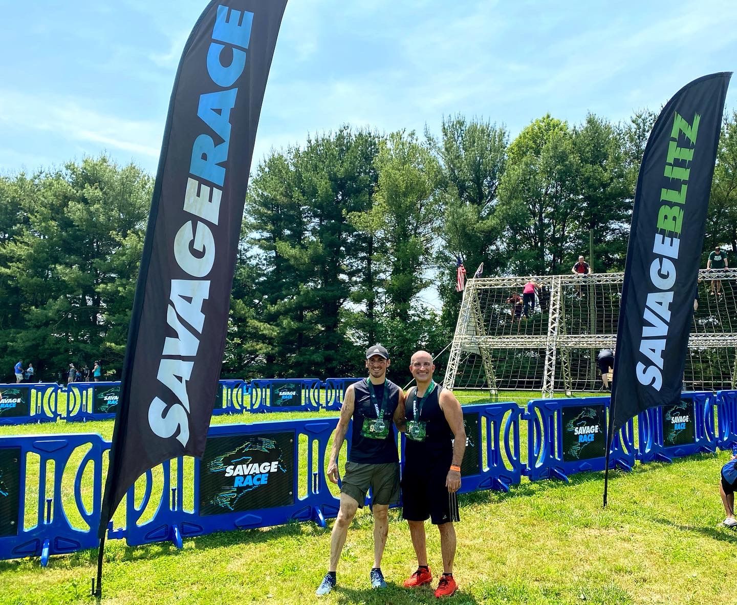 Two men at a Savage Race event, posing with flags and a course in the background, on a sunny day.