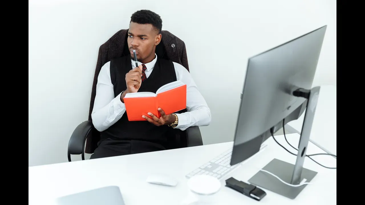 Man in vest, holding red notebook and pen, looks at computer screen, white desk, office setting.