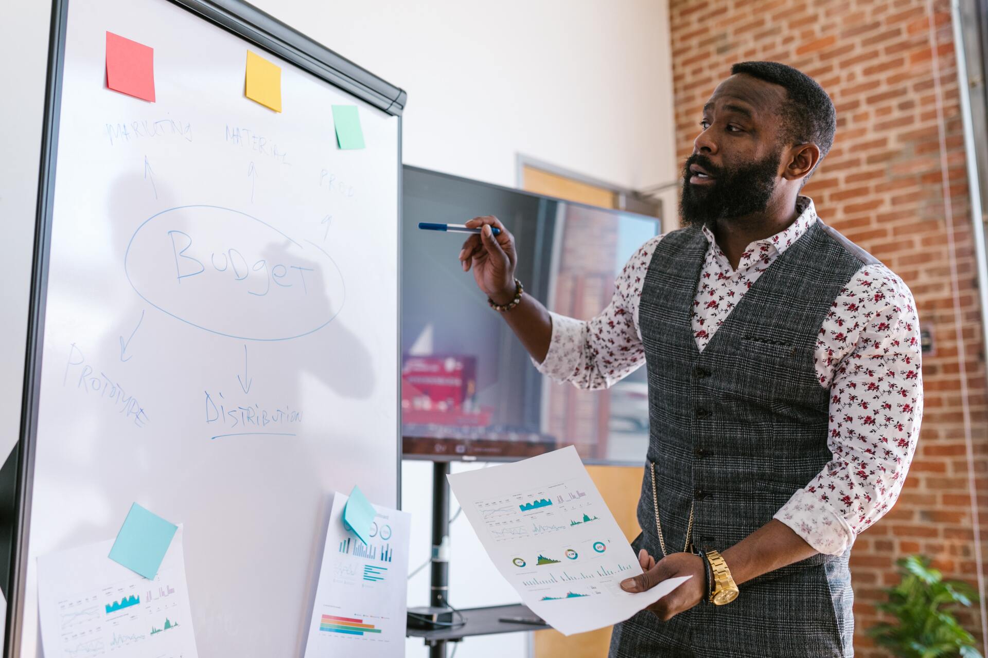 Man with a beard presenting at a whiteboard, holding papers, and wearing a vest in an office setting.