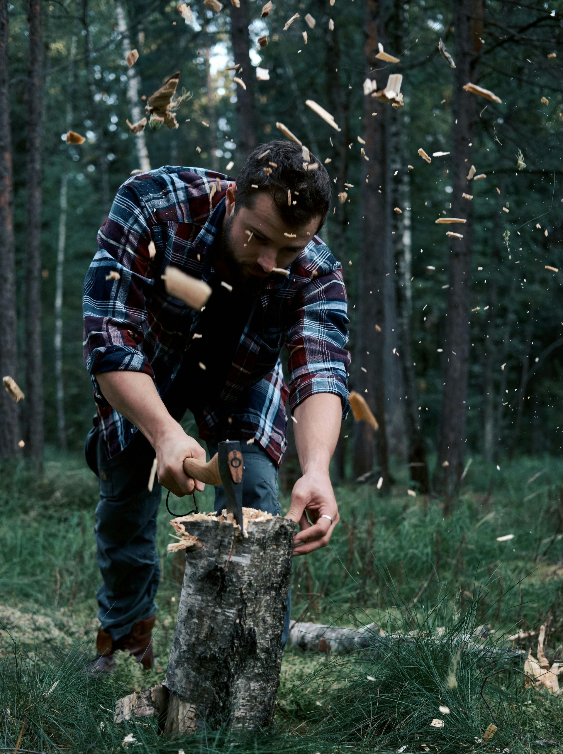 Man in flannel shirt chops wood with an ax in a forest, wood chips flying.
