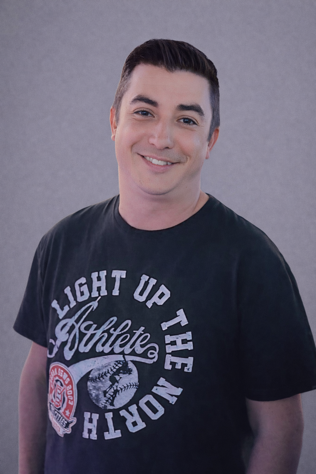 A headshot of Zac Douglas, a man smiling, wearing a black T-shirt, set against a gray background.